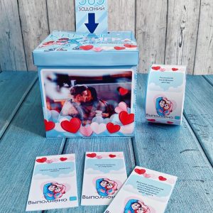A blue Valentine-themed game box set with hearts, a photo of a couple, and cards spread around it on a wooden surface; includes tasks and a completion card in Russian.