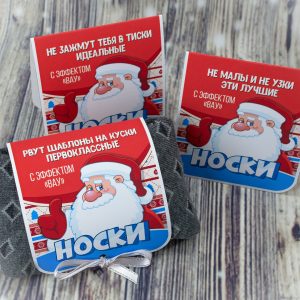 Red Christmas sock gift cards featuring Santa Claus and white text in Russian laid on a wooden surface with a gray fabric pouch nearby.